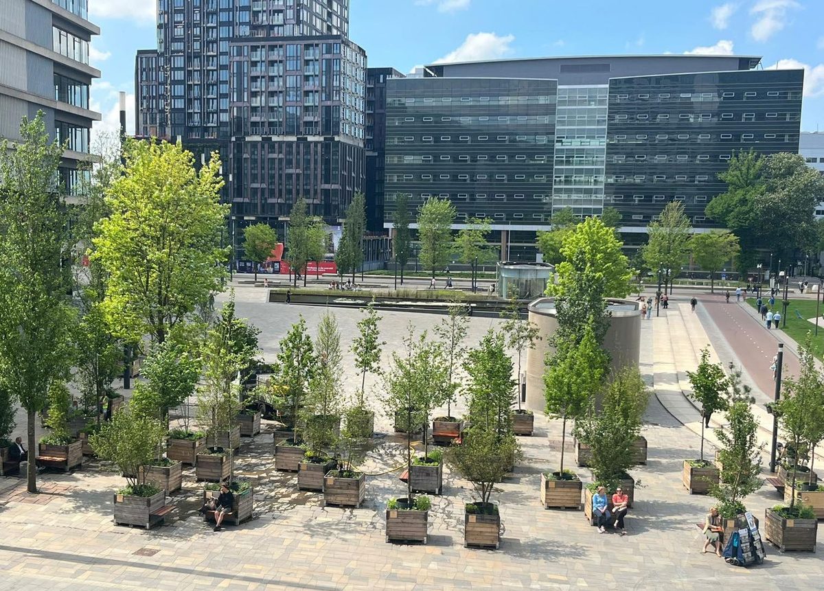 Stedelijk plein met groene schaduwmakers: verplaatsbare boomconstructies in plantenbakken tussen stadsgebouwen, met mensen die in de schaduw zitten.